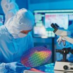 asian_male_technician_in_sterile_coverall_holds_wafer_that_reflects_many_different_colors_with_gloves_and_check_it_at_semiconductor_manufacturing_plant;_Shutterstock_ID_2110660535;_kostenstelle:_-;_job:_-;_sonstiges:_-